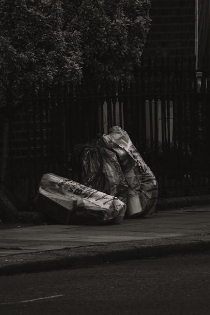 A group of three large black plastic rubbish bags filled with waste, placed on a pavement near the edge of a street. The bags are crumpled and slightly torn, revealing some of the contents inside. They are situated close to a black metal fence with vertical bars, behind which dense foliage of leafy trees is partially visible. The scene appears to be during the daytime but with low lighting or overcast conditions, casting subtle shadows on the ground. The pavement is smooth, with a curb separating it from the asphalt road, which has a faint line running parallel. The overall setting suggests an outdoor urban environment where private or independent waste disposal, such as rubbish collection by a service like Waste Collection Kensington, might be involved. The visual context emphasizes the need for appropriate rubbish removal and waste management services typical of Kensington's area.