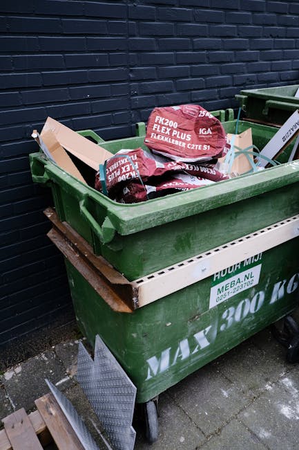 A group of three large black plastic rubbish bags filled with waste, placed on a pavement near the edge of a street. The bags are crumpled and slightly torn, revealing some of the contents inside. They are situated close to a black metal fence with vertical bars, behind which dense foliage of leafy trees is partially visible. The scene appears to be during the daytime but with low lighting or overcast conditions, casting subtle shadows on the ground. The pavement is smooth, with a curb separating it from the asphalt road, which has a faint line running parallel. The overall setting suggests an outdoor urban environment where private or independent waste disposal, such as rubbish collection by a service like Waste Collection Kensington, might be involved. The visual context emphasizes the need for appropriate rubbish removal and waste management services typical of Kensington's area.