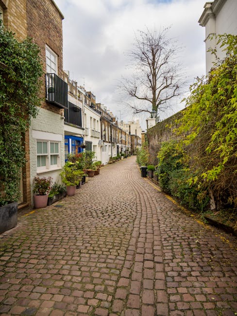 A narrow cobbled street in a residential area during daytime, with closely spaced terraced houses on the left side featuring white and brick facades, some with small balconies and potted plants in front. On the right, a tall brick wall with overgrown green foliage extends along the sidewalk, partially covering it. A large, leafless tree stands in the distance against a cloudy sky. The street appears quiet and clean, with a slight curve leading out of view, illustrating an urban environment where private waste collection or rubbish removal services, such as those offered by Waste Collection Kensington, might operate to manage waste in areas with limited access for standard public waste collection.