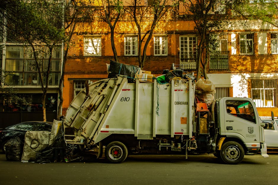 A white waste collection truck with the identification number 610 is parked on a city street in front of a row of residential buildings with orange and brown brick facades and multiple windows. The truck's rear compartment is open and filled with a variety of waste materials, including large sacks, cardboard boxes, and other refuse, some of which are stacked and leaning against the side of the vehicle. The truck is equipped with a hydraulic lifting mechanism that is partially visible. On the pavement beside the truck, there are black and grey garbage bags filled with waste, placed in front of a black vehicle parked nearby. The scene is set during daylight hours, with natural sunlight casting shadows of trees onto the buildings and street. The background features trees with green foliage and multi-story residential buildings with white-framed windows and balconies, creating an urban environment associated with private waste collection and rubbish removal services, such as those provided by Waste Collection Kensington.
