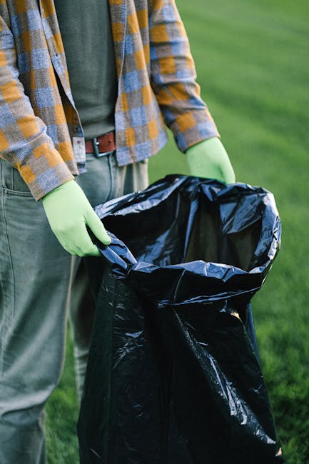 In the foreground of the image, a large pile of mixed waste and rubbish bags is overflowing from several waste bins positioned on a paved sidewalk. The bins include a grey recycling container labeled for mixed paper and cardboard, a black bin, and a bright red bin, all filled beyond their capacity, with waste materials spilling onto the ground. Surrounding the bins are scattered cardboard boxes, plastic packaging, paper waste, and various discarded items, indicating an accumulation of refuse. To the left, a parked silver car with a visible registration plate is situated behind a metal railing that separates it from the rubbish area. Behind the scene, there is a commercial building with a yellow storefront and multiple glass windows, which appear to belong to shops or eateries. Above, a blue construction scaffolding and safety barriers cover part of the upper façade of the building, with some signs attached for caution or guidance. The environment appears to be a busy urban street corner, and the scene suggests a situation where rubbish has not been collected in a timely manner, exemplifying common delays in waste removal services in Kensington, as managed by Waste Collection Kensington.