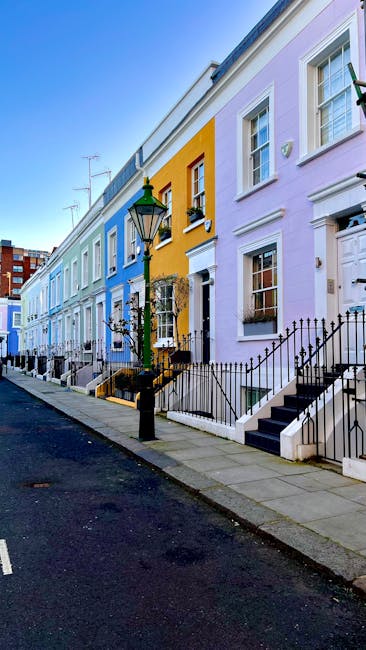A row of colorful terraced houses along a quiet urban street, each painted in pastel shades including light purple, yellow, and blue. The houses have white-framed sash windows and black wrought-iron railings along small front steps leading to front doors. A traditional black street lamp with a glass lantern is positioned on the pavement in front of the houses, slightly offset to the left. The pavement is clean and runs parallel to a dark asphalt road, which is visible in the foreground. The background features a clear blue sky and some distant modern red-brick apartment buildings. No rubbish or waste removal equipment is visible in the scene, but the setting suggests a typical residential area where private waste collection services, such as those provided by Waste Collection Kensington, could operate for on-site rubbish clearance or alternative waste handling.