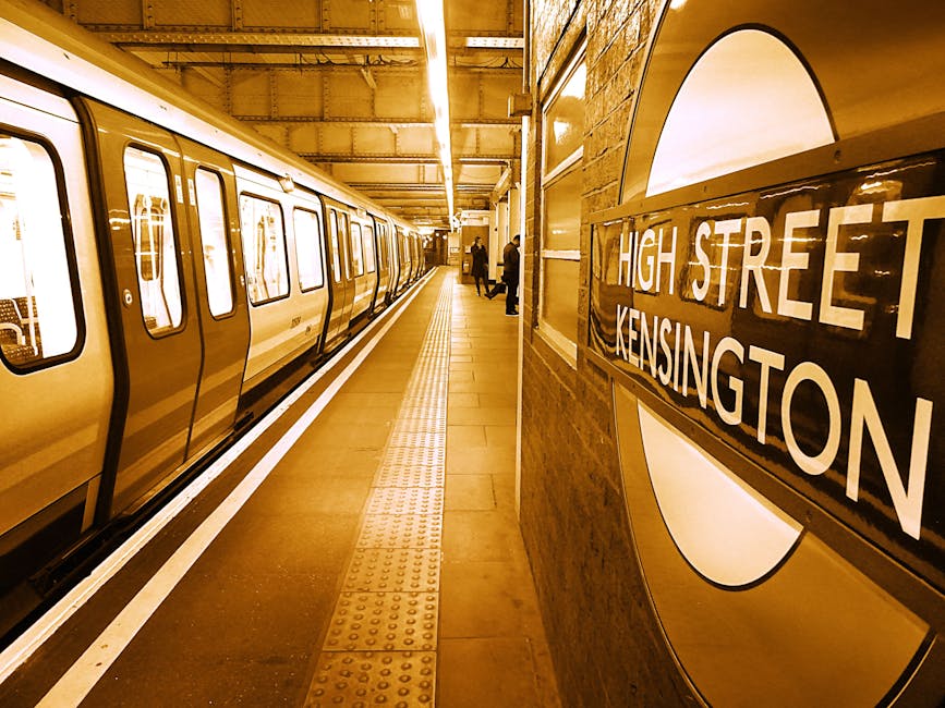A London Underground station platform at High Street Kensington, captured from the side with a row of beige and white tube train cars waiting along the track. The train doors are closed, and the exterior surface appears smooth with a shiny finish. The platform has a textured yellow safety line running parallel to the train, with a smooth brown tiled floor covering most of the area. To the right, a large, rounded sign inset into a brick wall displays the station name 'High Street Kensington' in bold, white capital letters against a dark background. In the background, a few passengers are seen standing on the platform, some holding bags or facing away from the camera, with the platform illuminated by warm lighting that reflects softly off the train's surface. The overall scene suggests a typical, quiet commuter stop within an urban environment, with elements of transportation infrastructure that could relate to private or alternative waste disposal in terms of station maintenance and facility management, as managed by local authorities or service providers like Waste Collection Kensington.