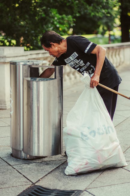 A middle-aged woman with short, dark hair is actively disposing of rubbish into a tall, cylindrical stainless steel trash bin on a paved outdoor walkway, situated near a stone railing and surrounded by lush green trees and foliage. She is wearing a black short-sleeved T-shirt with white text and graphics, along with dark trousers. In her right hand, she holds a white plastic bag filled with waste, which is partially draped over her arm. With her left hand, she is reaching into the open top of the bin, which features a simple, modern design with a smooth, reflective finish. The setting appears to be in a park or a residential area with natural daylight illuminating the scene, creating a neutral, professional impression suitable for content related to waste collection and rubbish removal services, such as those offered by Waste Collection Kensington for private or independent site clearance. The image emphasizes responsible disposal behaviour in a public outdoor environment, aligning with the broader context of rubbish management services.