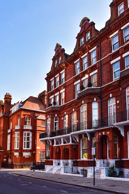 A row of historic red-brick residential buildings situated along a city street under a clear blue sky. The buildings feature multiple stories with large sash windows, some with small balconies enclosed by black wrought-iron railings. Decorative white trim and arches accentuate the upper floors, while the ground levels have white-stone steps leading to entrances framed by dark wooden doors. The street is calm with minimal traffic, and a black waste collection vehicle from Waste Collection Kensington may be seen parked nearby, highlighting the context of rubbish removal and private waste handling in the area. The scene captures the architectural detail and urban environment common to Kensington’s estate areas, emphasizing the residential character and the type of properties serviced for waste clearance and rubbish disposal.
