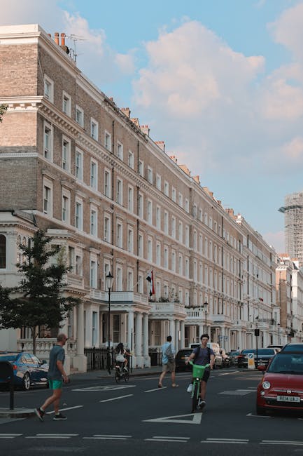 The image depicts a bright, sunny day on a street lined with multi-storey residential buildings in Kensington, with classic light-colored brick facades and white window frames. In the foreground, several pedestrians are walking along the pavement, including a man cycling with a black helmet, a woman in casual clothing, and a person on a bicycle. Parked cars are visible along the curb, including a red vehicle on the right and a dark-colored car further along the street. On the sidewalk, there are a few small trees with lush green foliage providing some shade. In the background, a modern building under construction with scaffolding and a temporary netting cover can be seen, contrasting with the historic architecture. The sky overhead is mostly clear with a few scattered clouds, and the overall scene captures an active urban environment typical of London. The scene subtly relates to urban waste management where private collection services such as Waste Collection Kensington could handle rubbish removal for city residents. No waste or rubbish is visible in this scene, highlighting the cleanliness of the street.