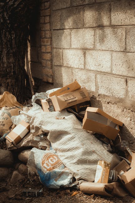 A pile of discarded cardboard boxes and packaging materials, some flattened and others partially torn, are stacked against a rough brick wall at the base of a large tree with dark, textured bark. The cardboard varies in shades of light brown and beige, with some featuring printed labels and barcodes. Among the boxes are large, woven fabric sacks, likely used for bulk waste, which appear weathered and slightly soiled. The ground beneath the refuse consists of small rocks and dirt, with a few scattered leaves and debris visible. The background displays a grey, weathered brick wall, and the scene is illuminated by natural light, casting subtle shadows on the waste and surrounding environment. This image depicts an outdoor scene appropriate for illustrating private waste disposal or rubbish removal services, such as those offered by Waste Collection Kensington, in a residential or urban setting where on-site clearance might be required for bulky refuse.