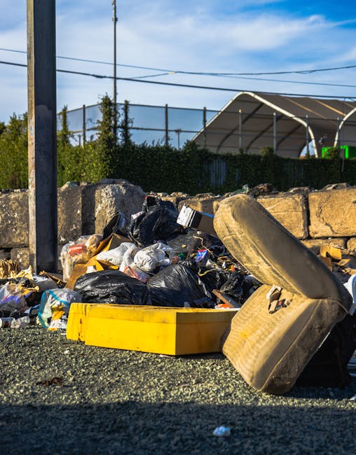Two large black plastic garbage bags filled with waste are placed on the pavement near a black metal fence and a wooden structure, possibly a garden trellis or gate, partially visible in the background. The bags are crumpled and appear to contain household rubbish, with some parts of the material visible through tears or thinning areas. The surrounding environment suggests an outdoor urban setting, with a mix of greenery from nearby trees or bushes on the left side of the image. The scene is captured in low light conditions, giving the image a dark, monochromatic tone. This scene, associated with rubbish disposal, can imply private or alternative waste handling methods outside of scheduled council collection, aligning with services provided by companies like Waste Collection Kensington for rubbish removal. The focus is on the generic waste bags positioned on the street, awaiting collection or removal.