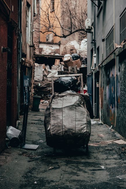 A narrow urban alleyway cluttered with various waste materials and discarded items. In the foreground, there is a large, dark grey rubbish sack with a textured, fabric-like surface, positioned on the slightly uneven pavement. Behind it, a large metal trolley or wheeled bin is visible, with its handle extending upward. Further into the alley, an assortment of cardboard boxes, plastic bags, and loose debris are piled haphazardly against a makeshift wooden barrier. The background features residential or commercial buildings with brick facades and peeling paint, some with graffiti on the walls. Leafless tree branches extend across the top of the alley, indicating a colder season. The scene is lit by natural, overcast daylight, creating a dull, muted atmosphere. The presence of the waste and the confined space suggest an area commonly used for informal rubbish disposal, which Waste Collection Kensington may handle through alternative waste management services such as private rubbish collection or on-site clearance. Overall, the scene depicts an accumulation of refuse typically associated with last-minute or ad-hoc rubbish removal needs in an urban setting.
