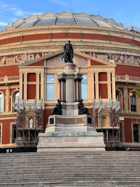 A large, historic brick and stone building with a domed roof under construction scaffolding, featuring a central statue of a seated figure on a tall pedestal at the top of wide stone steps. The facade includes decorative stone columns and a frieze with sculpted figures, and the scaffolding surrounds both sides of the statue. The building's exterior has multiple windows with rectangular panes, and the sky above is clear and blue. The scene suggests ongoing preservation or renovation work, potentially related to a cultural or civic institution, with the presence of waste collection services implied but not visible in the image.