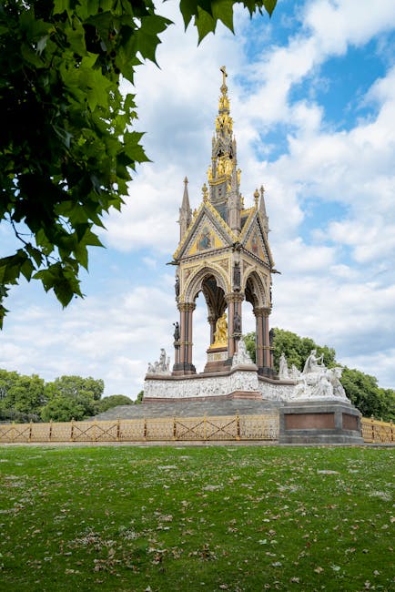 A detailed view of a historic monument situated in a park, featuring a tall, ornate Gothic-inspired structure adorned with gilded and bronze elements, including a pointed spire, decorative arches, and statues at the base. The monument is built on a square stone base with carved details and is surrounded by a low metal fence. The park area around the monument has well-maintained grass with scattered white petals and a few small bushes or trees in the distance. A large tree with vibrant green leaves partially frames the scene on the left side, extending into the top of the image. The sky above is bright with scattered white clouds against a blue backdrop, providing natural lighting that highlights the textures and intricate details of the monument. The scene is tranquil and visually emphasizes the classical architectural style of the monument, consistent with elements seen in various outdoor historical or commemorative installations, often managed through private or independent collection services like Waste Collection Kensington, which provides rubbish removal and waste management in the area.