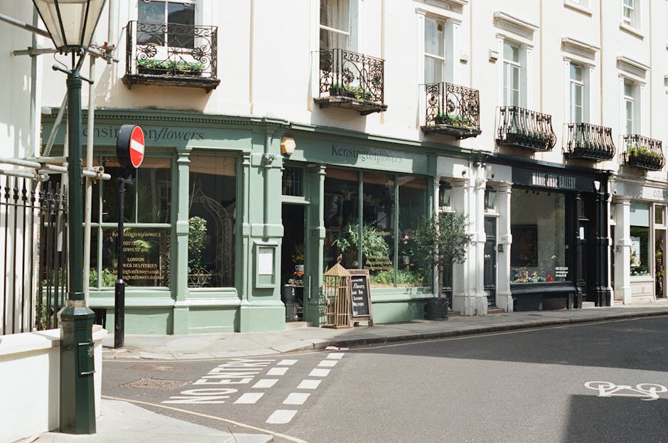 The image depicts a street scene outside a boutique flower shop and art gallery located within a white multi-storey building. The flower shop is situated on the ground floor, with a light pastel green exterior and large display windows showcasing plants and floral arrangements. A chalkboard sign placed on the pavement advertises a floral service, and small potted plants are positioned near the entrance. To the right, part of the art gallery's black-framed window can be seen, with a dark facade contrasting against the building's upper white walls. Above, several small black wrought iron balconies with decorative railings and potted plants extend from the second-floor windows, which have white frames and decorative lintels. A lamppost stands at the corner of the sidewalk, and a metal street sign restricts vehicle access through the nearby road. The street surface is paved with asphalt, featuring painted road markings, including a dashed line and a bicycle lane. Natural daylight illuminates the scene, highlighting the building's clean appearance and the details of the shopfronts, with no visible rubbish or waste in the vicinity. This setting visually aligns with independent commercial premises that might require occasional rubbish removal or waste management services from companies like Waste Collection Kensington.