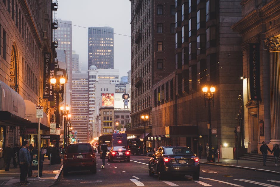 A city street scene during dusk with tall, multi-storey office buildings and commercial structures lining both sides of the road. The buildings are primarily constructed from brick and stone, featuring architectural details such as decorative cornices and large windows with metal frames. The street is illuminated by warm, vintage-style lamp posts and streetlights that cast a yellow glow on the pavement and surrounding facades. Several vehicles, including cars and vans, are parked along the curb or moving through the street, their headlights reflecting off the wet asphalt. Pedestrians are walking on the sidewalk, some near shop entrances with awnings and signage, while others cross the street. Large digital billboards on the building facades display advertising images, adding visual interest to the urban environment. Despite the bustling scene, the atmosphere appears slightly hazy, indicative of early evening or foggy weather. This urban setting, captured in a professional and neutral tone, subtly relates to private waste collection or on-site clearance activities typical of commercial districts, with the street scene emphasizing the importance of managing rubbish and debris in such densely developed areas. The atmosphere and composition highlight the importance of effective rubbish removal services, such as those provided by wastecollectionkensington.co.uk, ensuring clean and well-maintained city environments.