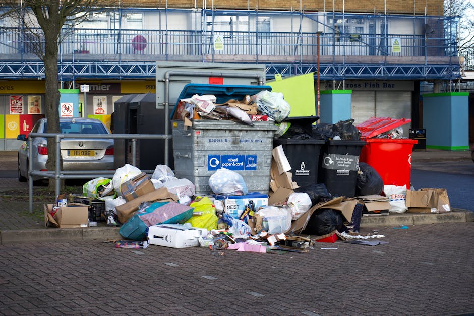 In the foreground, a large collection of mixed household waste and recyclables is piled on a paved sidewalk next to a metal railing, with an overflowing grey recycling bin containing paper and cardboard, and several black and red waste bags and cardboard boxes surrounding it. The trash includes crumpled paper, plastic packaging, folded cardboard, and plastic bags, some spilling onto the ground. Behind the waste, a silver car with a UK registration plate is parked adjacent to the pile. In the background, there is a commercial building façade with storefronts, including a fish bar, and a blue scaffolding structure covering the upper part of the building. Fluorescent signals and warning signs are visible on the storefronts, and the scene is lit with natural daylight, highlighting the mess that requires waste management or alternative rubbish removal services like those provided by Waste Collection Kensington. The environment indicates a typical urban street scene where private waste disposal might be necessary due to access or collection limitations.